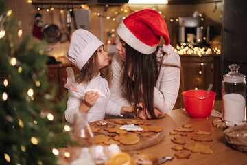 Happy little girl with her beautiful mom make gingerbread at home. Christmas decoration at kitchen. Fir tree with fairy lights