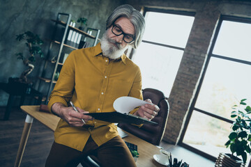 Photo of doubtful mature guy dressed yellow shirt spectacles reading contract before signing indoors workshop workstation