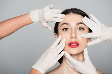 plastic surgeons in latex gloves touching face of brunette woman isolated on grey