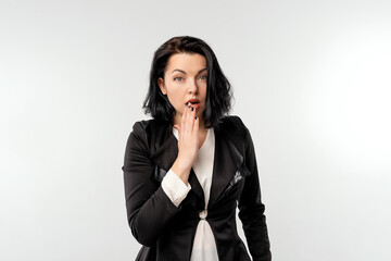 Coy cute young businesswoman in black formal jacket white shirt covering mouth and looking guilty, did something awkward, standing over white background