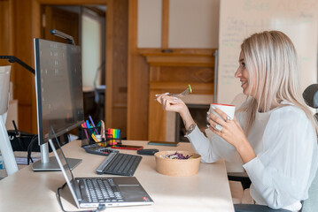 Beautiful young woman relaxing after work, eating lunch and drinking coffee by the computer.