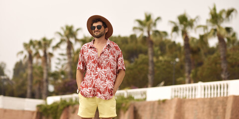 Tourist man on the beach standing outdoors against resort with palm trees in the background during summer holidays. The guy wearing a summer casual shirt hat and sunglasses, standing looking to the