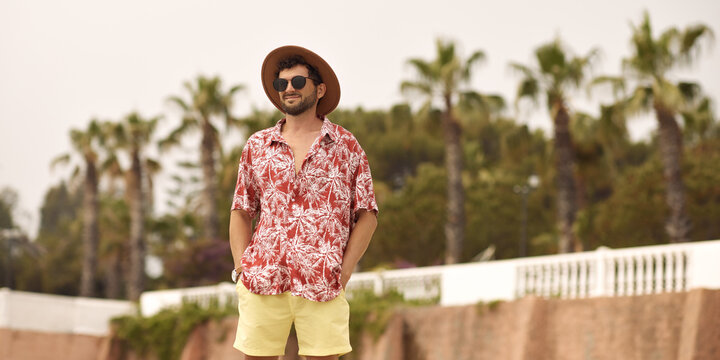 Tourist Man On The Beach Standing Outdoors Against Resort With Palm Trees In The Background During Summer Holidays. The Guy Wearing A Summer Casual Shirt Hat And Sunglasses, Standing Looking To The