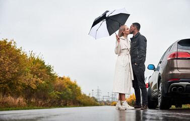 Young couple in love kissing under umbrella, standing on the road near cars under cloudy sky with rain. Low angle view