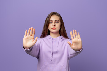 Serious young brunette woman showing stop gesture, saying no, prohibit something, wears purple...