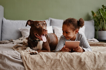 Front view portrait of cute African-American girl lying on bed with big pet dog and smiling, copy...