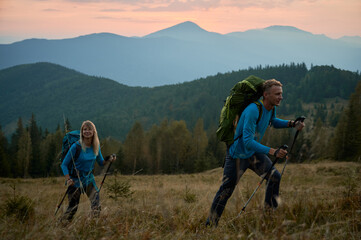 Obraz premium Young woman with glad smile on face following by her husband on hiking route while traveling on mountain spaces. Boundless woods and mountain ranges under the evening sky on the background.