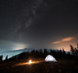 Fototapeta na wymiar Magnificent view of night starry sky with Milky way over grassy hill with illuminated camp tent and campfire. Camping during starry night. Coniferous forest and misty mountain peaks on the backdrop.
