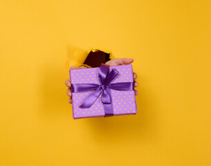 female hand holds a box with a gift on a yellow background, part of the body sticks out of a torn hole in a paper background. Congratulation, holiday surprise.
