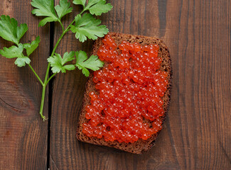 red salmon caviar lies on a piece of rye bread. Brown wooden table