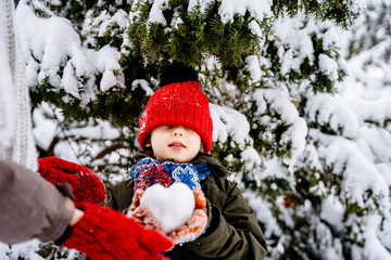 Unrecognizable mother giving snowball shape heart for her little son, dressed in colorful clothes are playing together with snowball in a winter park with snowy spruce on background.