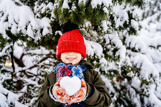 Funny little boy in warm winter clothes holding shape heart snowball with snowy fir trees on background. Outdoors winter activities for kids. Cute child wearing a warm red hat low over his eyes.