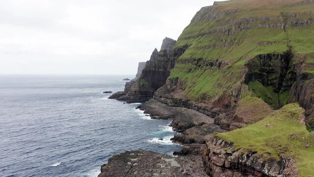 Aerial back view of huge cliffs in faroe islands, green rocky mountain,powerful ocean waves,in a cloudy summer day,green meadow and rock in wild nature,wild no house.