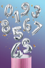 Shot of silver balloons. The balloons have are shaped as numbers from 0 to 9. The balloons are flying out of a pink box. The balloons are on the multicolored background.