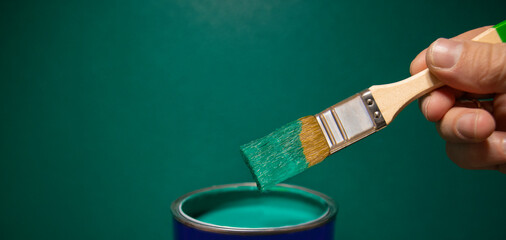 Man's hand holding a paintbrush over a can of paint on a green wall background, empty space for...