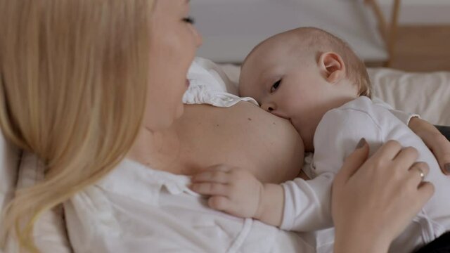 Young blonde woman feeding with breast and stroking her baby boy. Concept of breastfeeding, maternity and child healthcare. Time for togetherness, love and rest for mother and little son