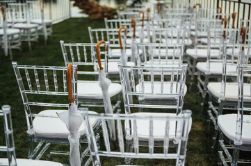 White glass chairs stand on the green grass. Round arch for a wedding ceremony in the background