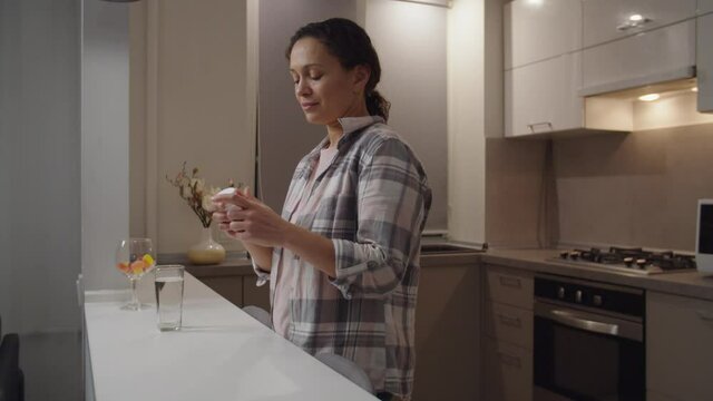 Beautiful lady taking daily medication or vitamin supplements for beauty at home. Adult woman holding slimming drug bottle, reading instruction, drinking glass of water to take diet pills indoors.