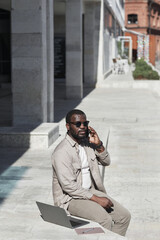 Vertical portrait of adult black man speaking by smartphone while working outdoors in city lit by sunlight