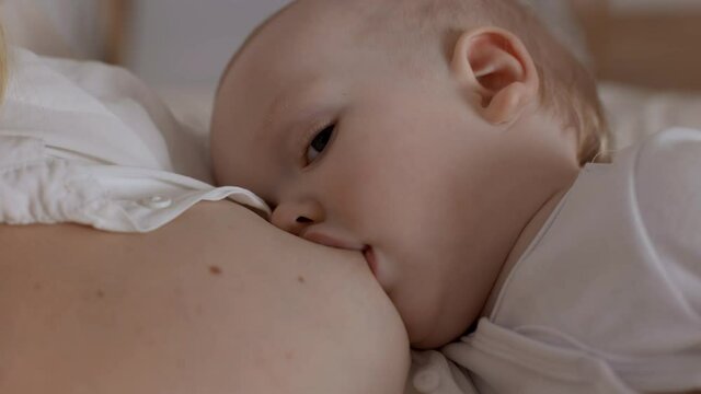 Baby boy drinking milk from mother breast and looking to camera. Concept of breastfeeding, maternity and healthy food for infant. Young woman feeding her hungry little son.