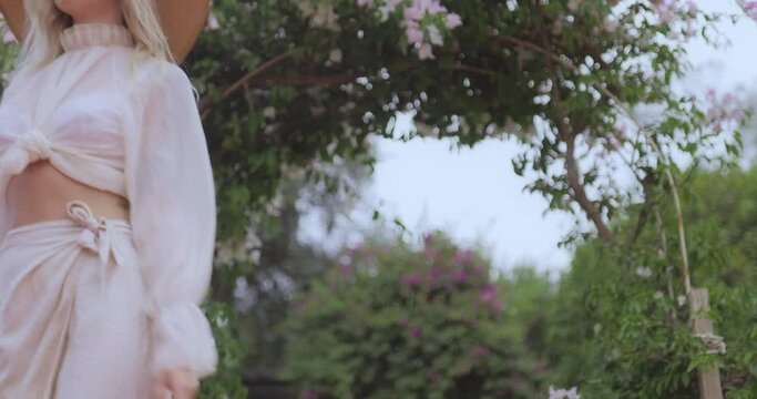 Romantic stylishly dressed young adult beautiful woman relaxing in a countryside park looking away. Blonde Parisian-styled model in a white transparent dress and straw hat walking against of garden