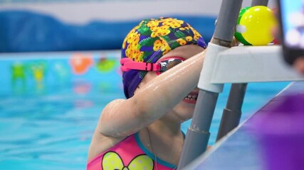 One young happy girl wear cap and pink goggles, smiling, holding a green ball in her hand, playing in blue water in the swimming pool. Close up portrait. Concept of active leisure with mom.