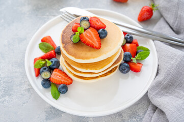 A stack of delicious fluffy pancakes with berries and honey on a white plate on a gray concrete background. Copy space.