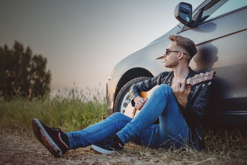 A man singer with the guitar is sitting by the car.