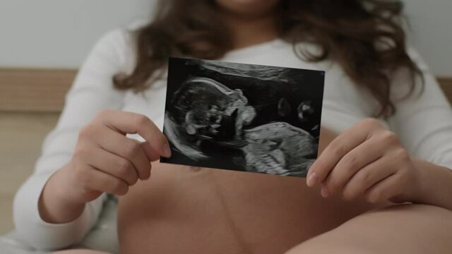 Expectant mother shows picture of unborn child to camera while lying in bed. Young pregnant woman holds photo of sonographic examination of baby in her belly