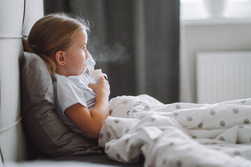 Little girl use inhaler nebulizer lying in bed in bedroom. Child asthma inhaler, nebulizer steam, flu or cold concept. Copyspace - obrazy, fototapety, plakaty