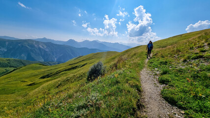 A man hiking along a narrow pathway in high Caucasus mountains in Georgia. There are high glaciers in the back. Thick clouds above the sharp peaks. Lush pastures on the sides. Barren peaks.