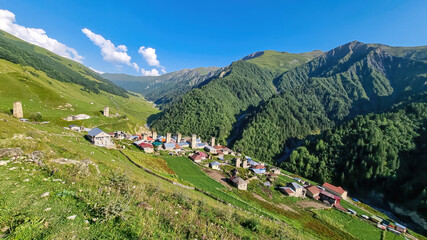 Fototapeta na wymiar Panoramic view on Adishi, a mountain village, located in high Caucasus mountain chains. Few building on the bottom of the valley. Lush green pastures with a few cows grazing on them. Idyllic landscape