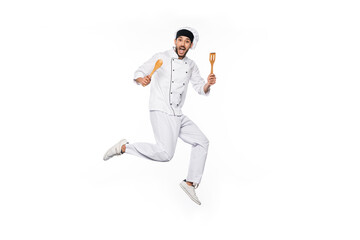 excited chef in hat and uniform jumping and holding wooden spatula and spoon isolated on white.