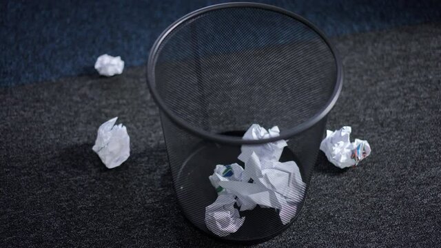 Black office trash bin with crumpled paper ball falling in slow motion inside. Unknown business person making mistake throwing worthless documents indoors. Failures and waste concept