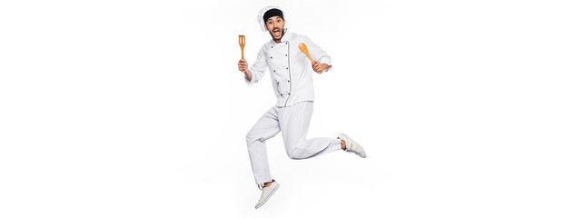 Fototapeta na wymiar excited chef in hat and uniform jumping and holding wooden spatula and spoon isolated on white, banner.
