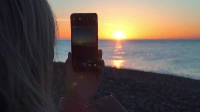 Women's hands hold smartphone and take pictures nature. tourist takes pictures with mobile phone camera of an amazing sunset on beach against background of sea or ocean. blonde girl in denim jacket.