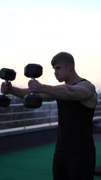 Caucasian white muscular man lifting dumbbells against the sunset sky background. Concept of desire, will and potential. 4k vertical footage 