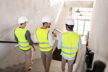 architecture, construction business and people concept - male architects in helmets with blueprints and clipboard walking downstairs at office building