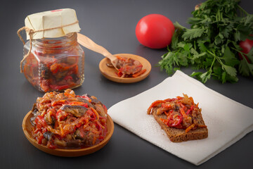 Stewed eggplant with vegetables in a wooden plate and a slice of bread with eggplant caviar on a white napkin on a gray table against the background of a glass jar, tomato and greens. Homemade. - obrazy, fototapety, plakaty