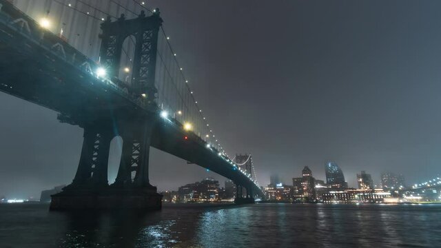 Zooming out timelapse of the illuminated Brooklyn bridge that crosses the East River in New York City, USA. Night. Vessels sail along the illuminated river, skyscrapers on the other bank