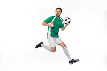 amazed football player in uniform jumping while holding soccer ball and showing thumb up on white.