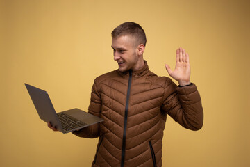 Young 20s man in brown jacket, on yellow background, holding a laptop computer using work,...