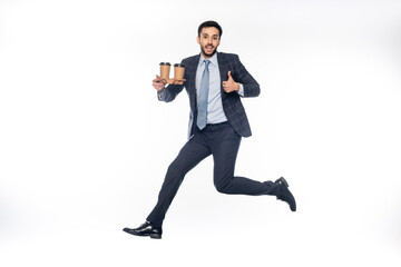 happy businessman in suit jumping while holding carton cup holder with paper cups and showing thumb up on white.