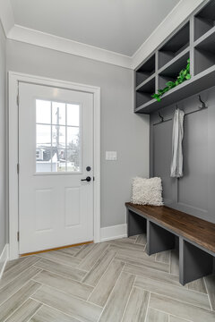 A Staged, Gray Mudroom / Entryway With Bench Seating, Coat Hooks, And Storage Above. A Scarf Hangs From A Hook And A Plant Sitting On A Shelf.	