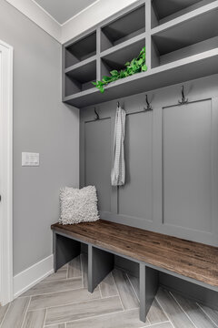 A Staged, Gray Mudroom / Entryway With Bench Seating, Coat Hooks, And Storage Above. A Scarf Hangs From A Hook And A Plant Sitting On A Shelf.