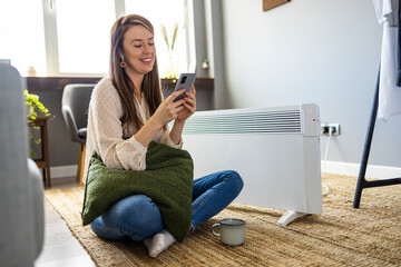 Woman dressed in beige sweater regulating heating temperature with a modern wireless thermostat and...