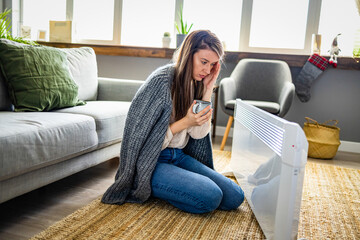 Obraz premium Young woman in woolen plaid warm up hands over electric heater, concept of loneliness, seasonal depression, stay home self isolation concept, coronavirus quarantine. The freezing girl about a heater