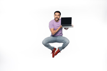 positive man in jeans and purple t-shirt levitating while holding laptop with blank screen on white.