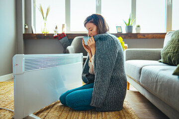 Young woman sitting near electric heater at home. Concept of heating season. Cold Woman In Jacket Sitting Near Heater At Home. Heavy duty radiator - central heating. Woman is getting her hands warm