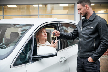 Naklejka na ściany i meble Handsome salesperson giving keys to young beautiful woman customer sitting in white car. Happy girl receiving key of her new automobile at auto dealership outdoors.
